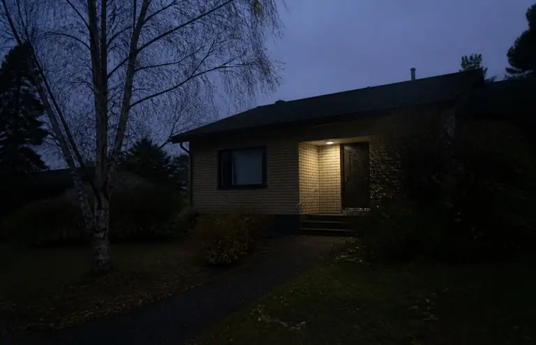 Omakotitalon etupiha iltahämärässä ennen: yksi kylmä kuistilamppu, kulkuväylä ja koivu pimeässä